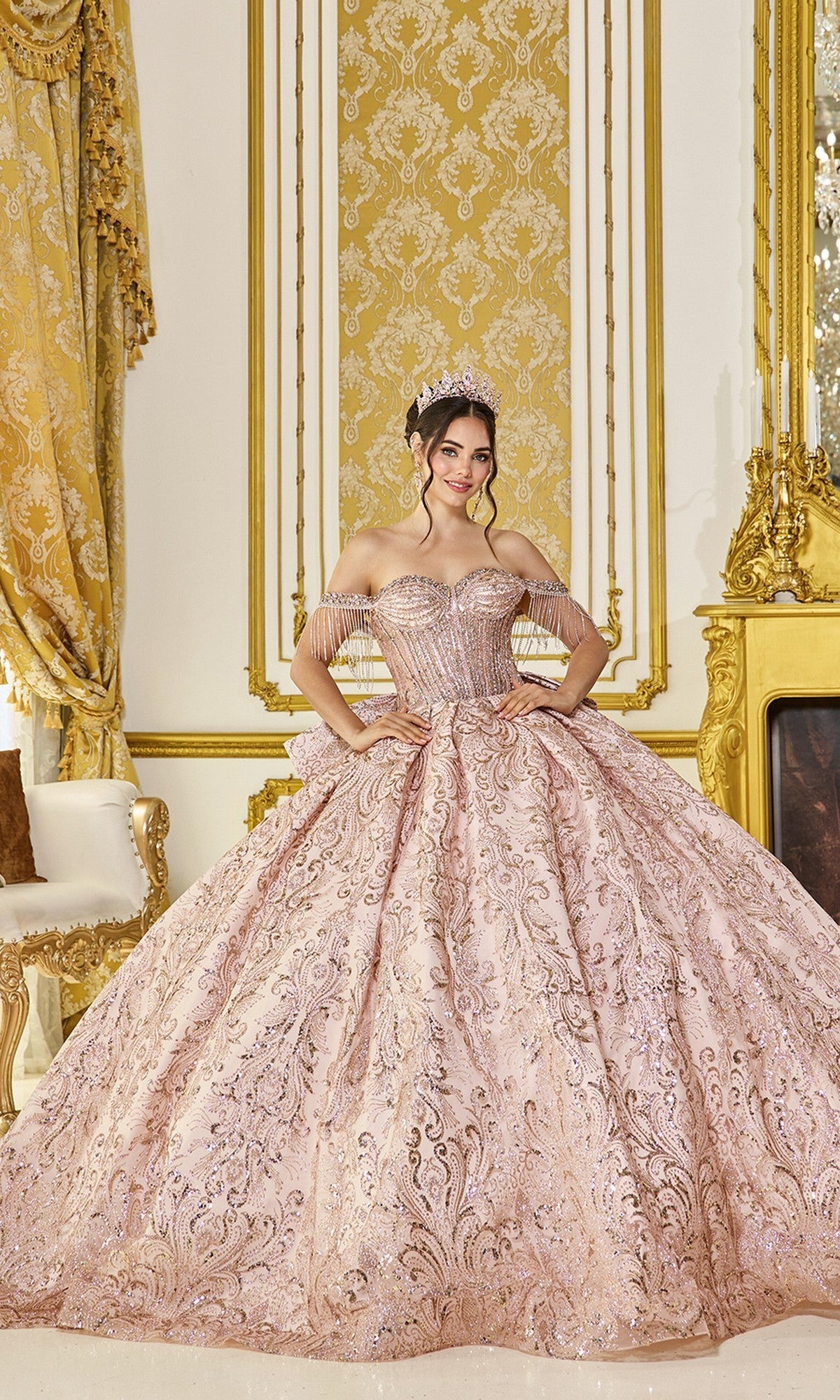 A young woman wears the Quinceanera Dress 5107 by Dancing Queen in shimmering rose gold and a tiara, standing in an ornate room with gold accents, patterned curtains, and a fireplace.